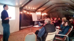 Man standing to the left of a projector screen speaking to a group of people sitting in a tent.