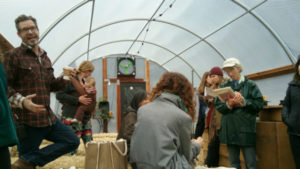 A group of people standing in a hoop house-style greenhouse with one man speaking and other taking notes
